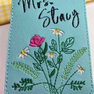 May include: A light blue luggage tag with the name "Mrs. Stacy" in black script. The tag features an embroidered floral design with a pink rose, white daisies, and green leaves. The tag has a stitched border and a small handle.