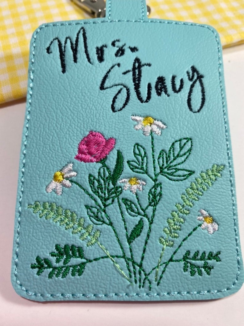 May include: A light blue luggage tag with the name "Mrs. Stacy" in black script. The tag features an embroidered floral design with a pink rose, white daisies, and green leaves. The tag has a stitched border and a small handle.
