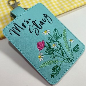 May include: A light blue rectangular keychain with the embroidered text "Mrs. Stacy" in black script. The keychain features a floral design with a pink rose, white daisies, and green leaves. It has a silver-colored clasp.