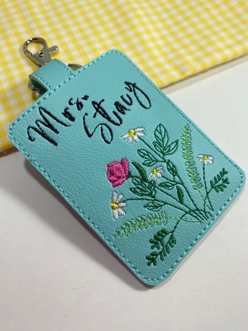 May include: A light blue rectangular keychain with the embroidered text "Mrs. Stacy" in black script. The keychain features a floral design with a pink rose, white daisies, and green leaves. It has a silver-colored clasp.