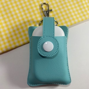 May include: A teal leatherette AirPod case with a silver clasp. The case has a snap closure with a white button. The background is a yellow and white checkered pattern.
