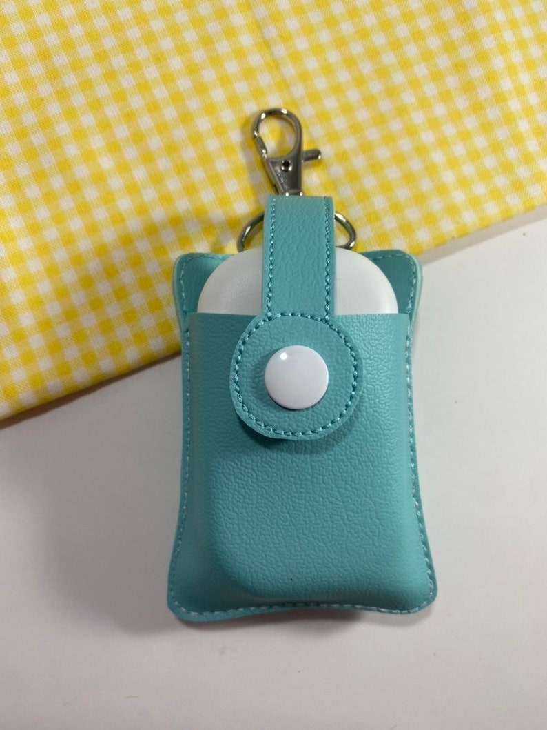 May include: A teal leatherette AirPod case with a silver clasp. The case has a snap closure with a white button. The background is a yellow and white checkered pattern.