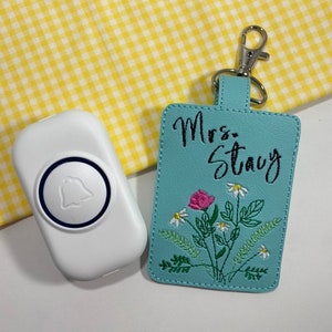 May include: A white doorbell with a blue bell icon next to a light blue keychain. The keychain has the name "Mrs. Stacy" embroidered above a floral design with a pink rose and white daisies. A silver clasp is attached.