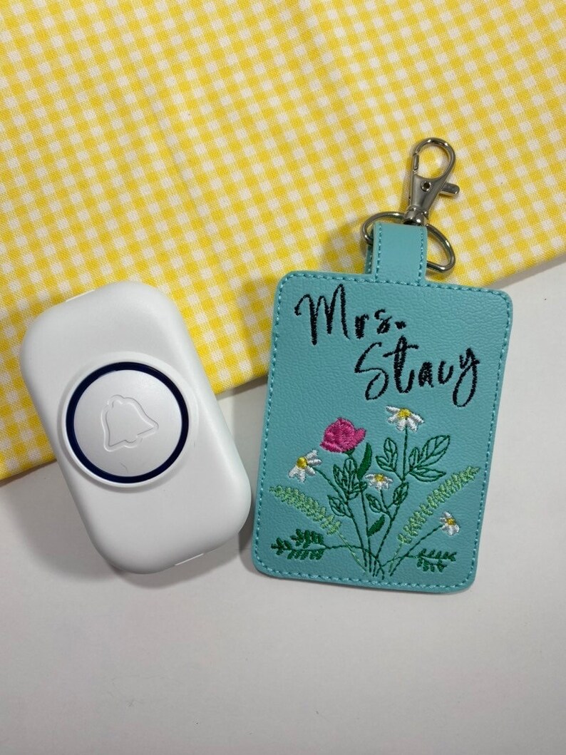 May include: A white doorbell with a blue bell icon next to a light blue keychain. The keychain has the name "Mrs. Stacy" embroidered above a floral design with a pink rose and white daisies. A silver clasp is attached.