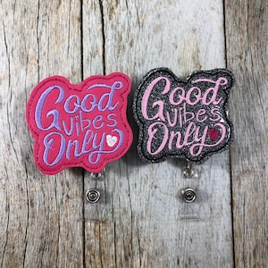 Peut inclure: Deux porte-badges avec l'inscription « Good Vibes Only ». L'un est rose avec des lettres violettes et un cœur blanc. L'autre est noir avec des lettres pailletées roses et un cœur rouge. Les deux ont un cordon rétractable et un clip en métal.
