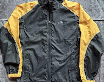 May include: A charcoal gray and mustard yellow windbreaker jacket featuring a full-length front zipper, a high collar, and long sleeves with elastic cuffs. The North Face logo is embroidered in white on the upper left chest area.