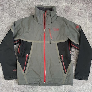 Pode incluir: Casaco cinza e preto para atividades ao ar livre com zíper e detalhes em vermelho. O casaco tem gola alta e vários bolsos com zíperes vermelhos. As mangas são pretas e o logotipo "Mountain Hardwear" é vermelho.