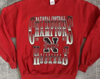 Peut inclure: Sweat-shirt rouge avec l'inscription "NATIONAL FOOTBALL CHAMPIONS 1997" en argent et noir. Le sweat-shirt présente également le logo "N" du Nebraska et le mot "Huskers".