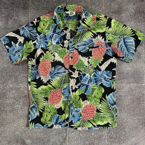 Peut inclure: Chemise à manches courtes à imprimé tropical. Le chemisier présente un fond noir avec un motif d'ananas rouges, de feuilles bleues et vertes et des accents blancs. Le col comporte une petite étiquette avec du texte.