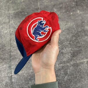 May include: A red baseball cap with a blue brim and a blue embroidered bear logo inside a white letter C. The cap features a lightweight, slightly crinkled fabric texture and a structured crown design, showcasing the iconic sports team emblem.