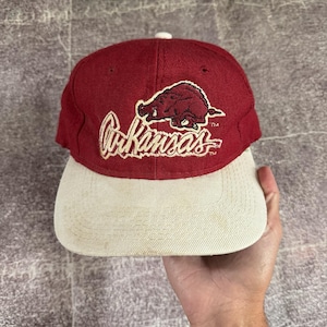 Peut inclure: Une casquette de baseball marron avec une visière couleur crème. L'avant présente un motif brodé d'un sanglier au-dessus du mot Arkansas écrit en lettres cursives. La broderie est de couleur crème avec un fin contour marron.