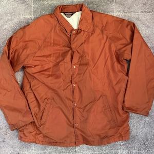 May include: A burnt orange coach jacket featuring a pointed collar, long sleeves, and a front button-down closure with matching orange buttons. The garment includes two side pockets and a light-colored interior lining with a KingSport brand label.