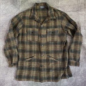 Puede incluir: Una camisa a cuadros marrón y verde con dos bolsillos en el pecho y dos bolsillos inferiores. La camisa tiene cuello y puños abotonados. La tela parece ser un material grueso y tejido.