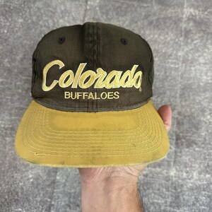 Könnte beinhalten: Vintage Baseballcap mit brauner Krone und gelbem Schirm. Das Wort "Colorado" ist in goldener Schrift über "BUFFALOES" gestickt. Der Hut ist ein klassischer Stil, wahrscheinlich aus den 1990er oder frühen 2000er Jahren.