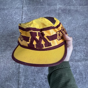 May include: A yellow and maroon cap with a curved brim. The cap features the letter "N" in maroon, with additional maroon accents and text. The cap is made of a durable fabric and has a structured design.