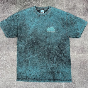 May include: Teal and black acid wash t-shirt with a crew neck and short sleeves. The shirt features a small logo on the upper left chest that reads "Lava Surfers" in a stylized font. The fabric has a mottled, textured appearance.