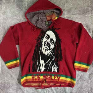 Puede incluir: Sudadera roja con cremallera y un retrato en blanco y negro de Bob Marley. La sudadera tiene un interior a rayas, un bolsillo delantero y puños y dobladillo a rayas de colores. El nombre "BOB MARLEY" está impreso en la parte inferior.