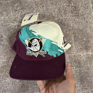 May include: A baseball cap featuring a maroon brim and lower section, a cream-coloured upper section, and a teal brushstroke graphic. The front displays an embroidered duck goalie mask logo with crossed hockey sticks. A tag is attached to the top.