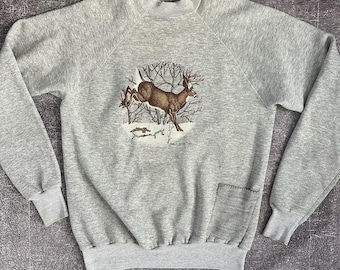 May include: A gray sweatshirt with a deer print on the front. The deer is jumping over a fallen log in a snowy forest. The sweatshirt has a pocket on the left side.