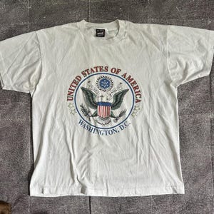 Puede incluir: Camiseta de color crema con un gráfico circular. El gráfico presenta las palabras "UNITED STATES OF AMERICA" y "WASHINGTON, D.C." alrededor de un emblema de águila. El emblema incluye un escudo con rayas rojas y blancas y un patrón de estrellas azules.