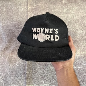Op de afbeelding: Een zwarte honkbalpet met de witte tekst WAYNE'S WORLD op de voorkant. De letter O in het woord WORLD is vervangen door een afbeelding van een wereldbol met zichtbare rasterlijnen. De pet heeft een gestructureerde voorzijde.