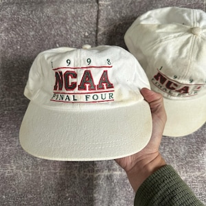 Puede incluir: Dos gorras de béisbol blancas con viseras curvas. Las gorras tienen el texto "1998 NCAA FINAL FOUR" bordado en rojo y verde. Las gorras están ligeramente desgastadas.