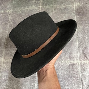 May include: A black fedora hat with a flat crown and a wide brim. The hat features a thin, smooth brown leather band wrapped around the base of the crown. The material of the hat appears to have a soft, felt-like texture throughout the surface.