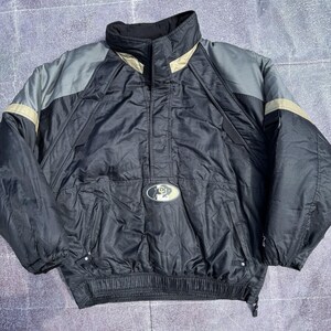 May include: A black pullover jacket featuring grey shoulder panels and tan accents. The jacket has a high collar, a central front pocket with a small oval logo patch, long sleeves with elastic cuffs, and a drawstring hem for an adjustable fit.