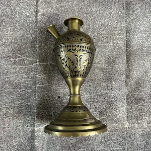 May include: An antique brass hookah with intricate black etched floral and fish designs. The hookah features a rounded body, a slender stem, and a flared base. The mouthpiece and top are also brass.