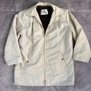 May include: A beige zip-up jacket featuring a dark brown faux fur lining, a pointed collar, and two front pockets with snap closures. The interior label displays the text BLAIR. The jacket has long sleeves and a straight hemline.