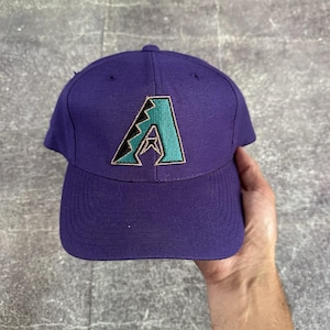 May include: A purple baseball cap featuring an embroidered teal and black letter "A". The cap has a curved peak and a button on top. The cap is made of a durable fabric.