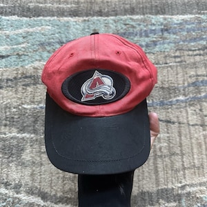 May include: Red and black baseball cap with a black and white embroidered logo of the Colorado Avalanche hockey team. The logo features the letter "A" with a mountain range and a hockey stick.