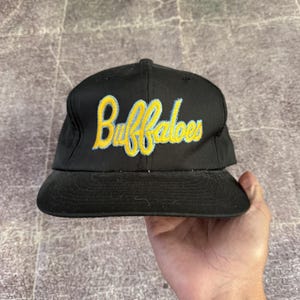 Peut inclure: Une casquette de baseball noire avec le mot Buffaloes brodé en lettres cursives jaunes et un contour bleu clair sur le panneau avant. La casquette présente une calotte structurée, une visière plate et des œillets d'aération.