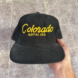 Peut inclure: Une casquette de baseball noire avec le mot Colorado écrit en lettres cursives jaunes au-dessus du mot BUFFALOES en lettres capitales jaunes. La casquette présente une calotte structurée et une visière plate en tissu noir.