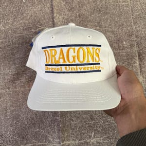Può includere: Un cappellino da baseball bianco con testo ricamato che recita DRAGONS in grandi lettere gialle con bordi blu, e Drexel University in lettere gialle più piccole sottostanti. Il cappellino presenta una corona strutturata e una visiera piatta.