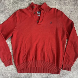 May include: A red knitted sweater with a ribbed collar and cuffs. The sweater features a button-up neckline and a small embroidered logo on the chest. The garment is made of a soft knit material.