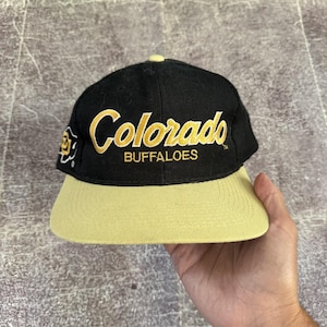 Op de afbeelding: Een zwarte honkbalpet met een platte gouden klep en een gouden knoop aan de bovenkant. De voorkant is voorzien van de geborduurde tekst Colorado BUFFALOES in goud, en de zijkant toont een klein wit en goud geborduurd buffellogo.