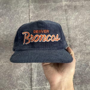 Puede incluir: Gorra de béisbol de pana azul marino con la palabra "Denver" sobre la palabra "Broncos" en bordado naranja. La gorra tiene una visera curva y una corona estructurada. Un accesorio deportivo clásico.