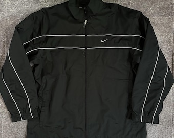 Puede incluir: Chaqueta deportiva negra de Nike con rayas blancas en el pecho y en las mangas. Cuenta con cremallera completa, un pequeño logotipo de Nike y cuello alto. La chaqueta tiene un cordón en la parte inferior.