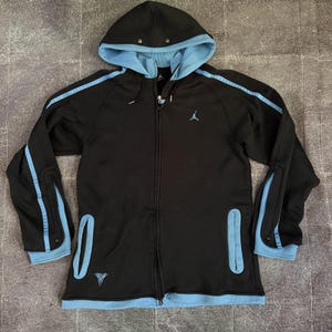 May include: A black zip-up hoodie featuring light blue accents along the sleeves, hem, and hood lining. The garment includes a front zipper, two vertical pockets, and a light blue Jumpman logo on the chest with a small graphic near the hem.