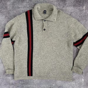 May include: A light gray knit sweater featuring a fold-over collar with two buttons at the neckline. The garment displays a vertical black and red stripe pattern on the right side of the chest and matching horizontal stripes on both sleeves.