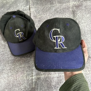 May include: Two black baseball caps with a purple bill. The caps feature the embroidered letters "CR" in white and purple. The caps are likely made of a durable fabric, suitable for outdoor wear.