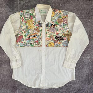 May include: White button-down shirt with a colorful patchwork design on the chest and collar. The patchwork features various graphics, including text, flowers, and cartoon characters. The shirt has long sleeves and a classic collar.