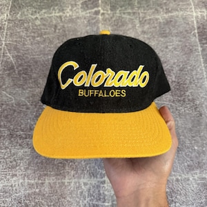 Op de afbeelding: Een zwarte honkbalpet met een gele klep en een gele knoop aan de bovenkant. Op de voorkant staat het woord Colorado in een geel cursief lettertype, met het woord BUFFALOES in gele hoofdletters direct daaronder.