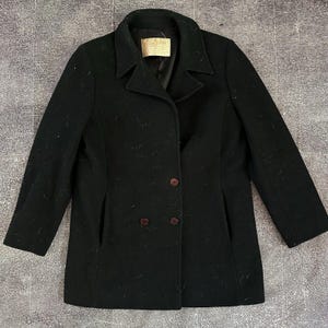 May include: A black wool coat with a double-breasted design. The coat features a notched collar, two rows of dark brown buttons, and two front pockets. A small label is visible near the collar. The coat is a classic outerwear piece.
