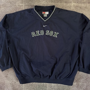 May include: A navy blue Nike long-sleeve pullover windbreaker featuring a V-neck collar with white trim. The chest displays the text RED SOX in dark green lettering with white outlines, positioned below a small white Nike swoosh logo.