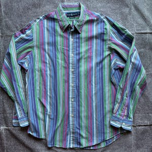 May include: A long-sleeve button-down shirt featuring a vertical striped pattern in shades of blue, green, purple, and white. The shirt has a pointed collar, a front button placket, and a small embroidered logo on the left chest area.