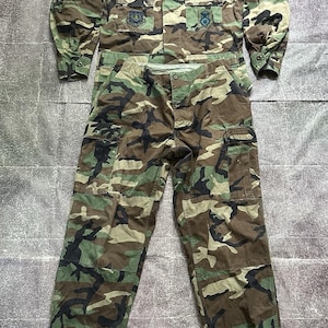 May include: A camouflage uniform set consisting of a long-sleeved button-up shirt and matching cargo pants. The shirt features a collar, two chest pockets, and patches including the text US AIR FORCE. The pants have side cargo pockets.