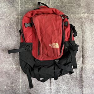 以下が含まれることがあります： 前面にゴールドの文字で「The North Face」と記された赤と黒のバックパック。複数の黒い調節可能なストラップ、サイドバックル、大きなジッパー付きコンパートメントを備えており、耐久性のある生地で構成されています。