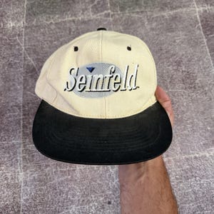 Peut inclure: Une casquette de baseball couleur crème avec une visière noire. L'avant présente un écusson ovale brodé en gris clair avec le mot Seinfeld en lettres cursives noires et une petite icône de triangle bleu au-dessus de la lettre i.